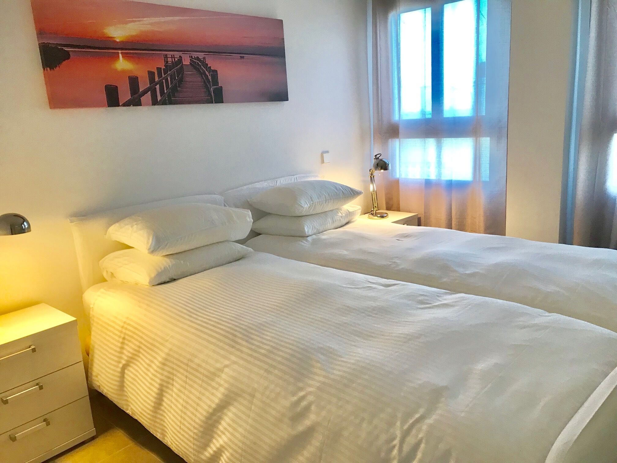 Una habitación de hotel con dos camas, una mesita de noche con lámpara, un cuadro en la pared y una ventana con cortinas.