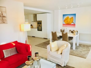 Una sala moderna con un sofá rojo, un sillón blanco y una mesa de centro de vidrio. Se ve un comedor con una mesa y sillas blancas al fondo.
