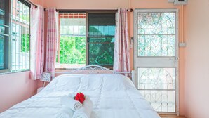 Una cama bien hecha con toallas enrolladas y una rosa, una ventana con vistas a follaje verde, y una puerta con un panel de vidrio decorativo.