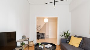 Een moderne woonkamer met een flatscreen televisie, een bank met een geel kussen, een salontafel met planten en een eethoek op de achtergrond.