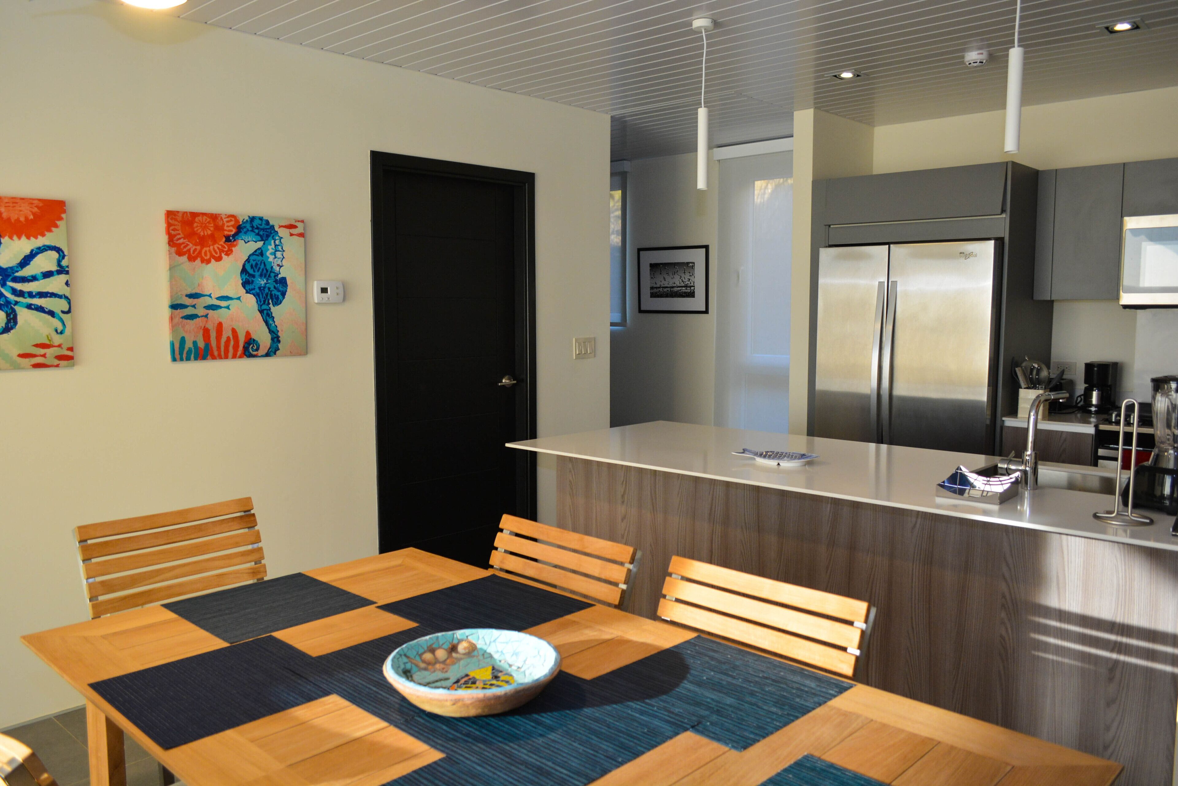 A modern kitchen with a built-in refrigerator, wooden dining table with chairs, and two wall-mounted paintings.