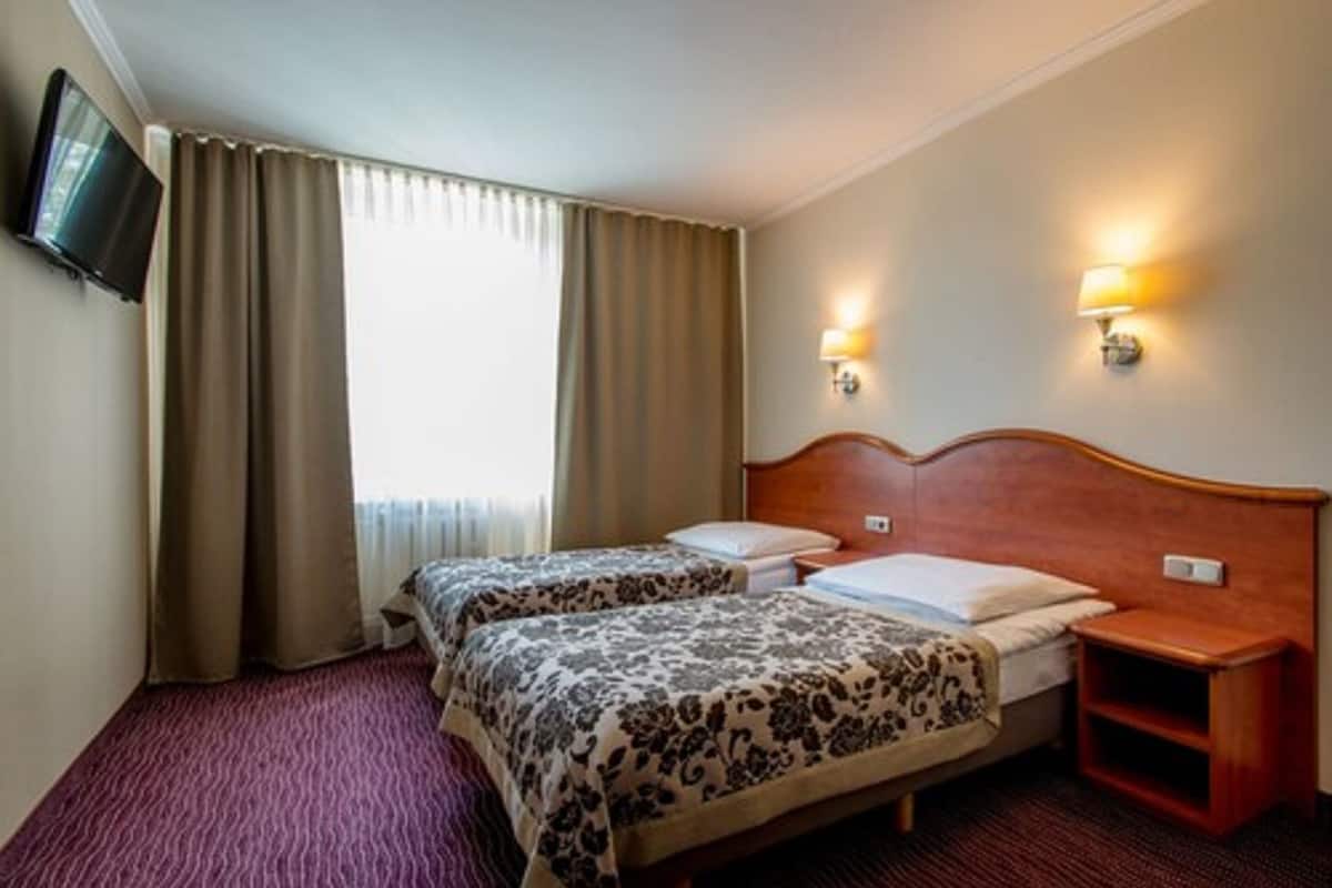 A hotel room with two beds, a TV mounted on the wall, and two wall-mounted lamps.
