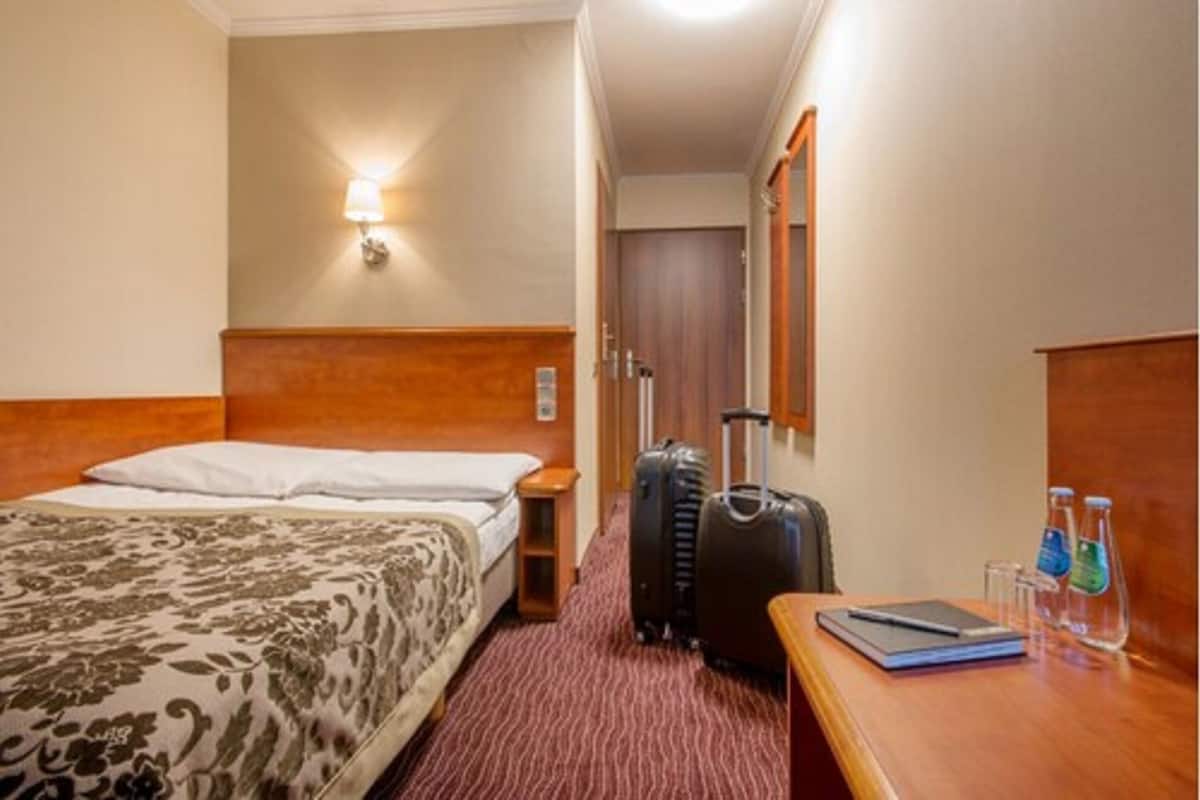 A hotel room with a bed, a nightstand, a desk with a laptop, two suitcases, and a wall-mounted lamp.