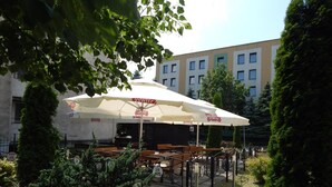 An outdoor seating area with tables and umbrellas, surrounded by greenery and a building in the background. - Hotel Krakus (Kraków)
