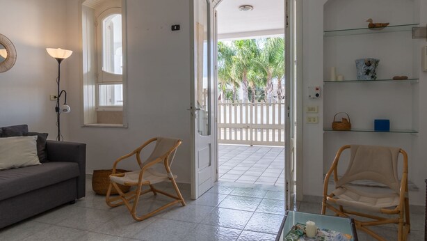 Una habitación con un sofá gris, dos sillones de mimbre, una mesa de centro de vidrio y una puerta blanca que da a un espacio exterior con valla blanca y palmeras.