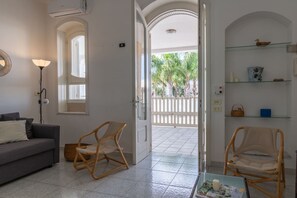 Een kamer met een grijze bank, twee rieten stoelen, een glazen salontafel, en een witte deur die leidt naar een buitenruimte met een wit hek en palmbomen.