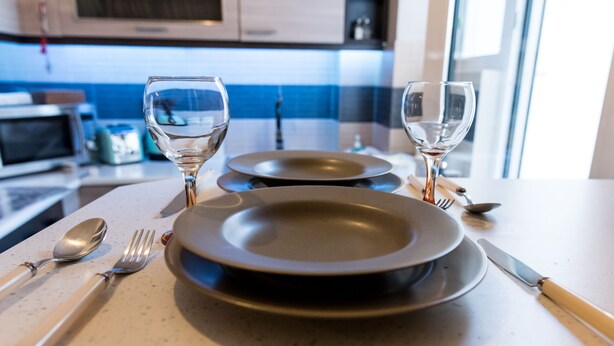 Een eettafel gedekt met borden, bestek en glazen, met een keuken op de achtergrond.