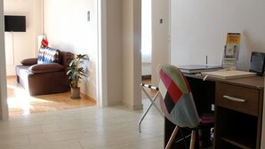 Een ruime woonkamer met houten vloer, een bruine bank, een kleurrijke fauteuil, een bureau met computer en een wandgemonteerde televisie.