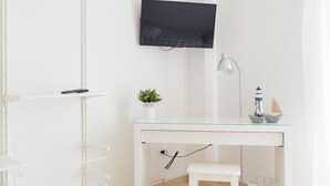 Een witte kamer met een glazen bureau, een wandgemonteerde televisie, een kleine potplant, een bureaulamp en een model schip.