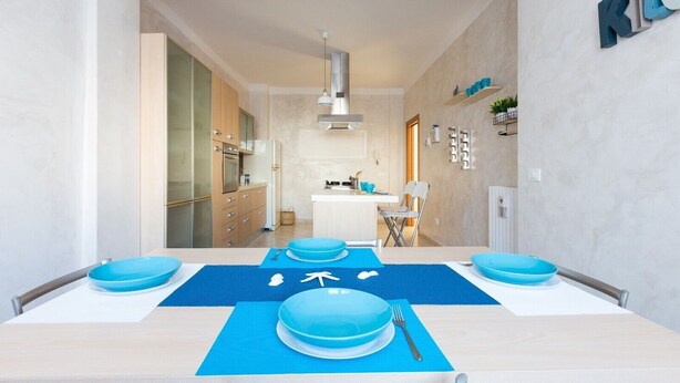 Een moderne keuken met een centraal eiland, een eettafel gedekt met blauwe borden en een blauwe placemat, en een keukenblad met een spoelbak en kastjes.