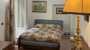Un dormitorio con cama, mesita de noche, lámpara, ventana con cortinas e imagen colgada en la pared.