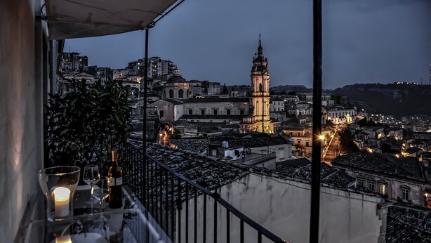 A balcony with a table set for two, overlooking a cityscape at dusk with illuminated buildings and a prominent tower. - Charming artist's hideaway with stunning view over Modica (modica)