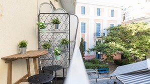 Um espaço exterior estreito com um portão de metal, uma mesa de madeira, dois taburetes pretos e vasos suspensos com vegetação.