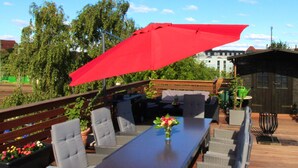 Une terrasse extérieure avec une longue table, plusieurs chaises et un parasol rouge.
