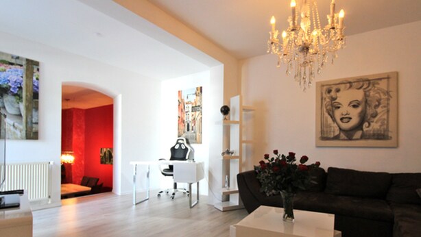 Un salon moderne avec un canapé foncé, une table basse blanche et un lustre. Un tableau orne le mur, et un mur d’accent rouge se trouve à l’arrière-plan.