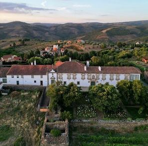 A large white building with a red-tiled roof, surrounded by greenery and set against a hilly landscape.