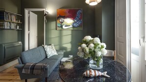 Ein Wohnzimmer mit einer Couch, einem runden Tisch mit Blumenvase, einem roten Sessel und einem Bücherregal.