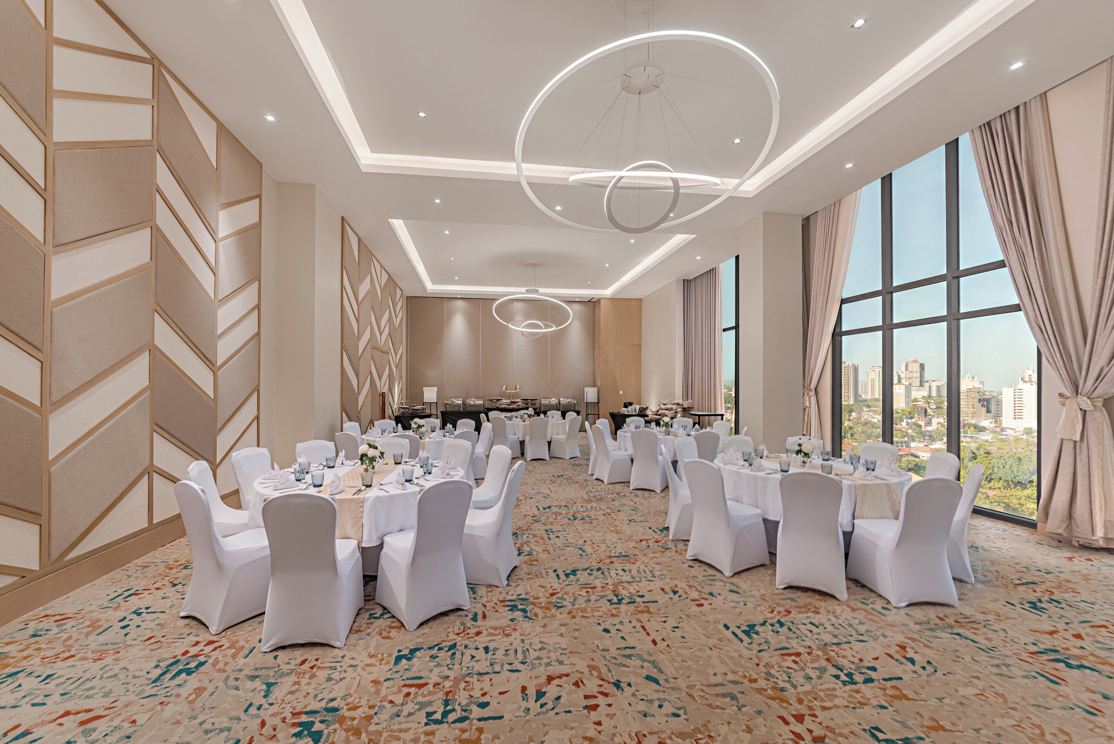 A banquet hall with round tables, white chairs, and a large window with a city view.