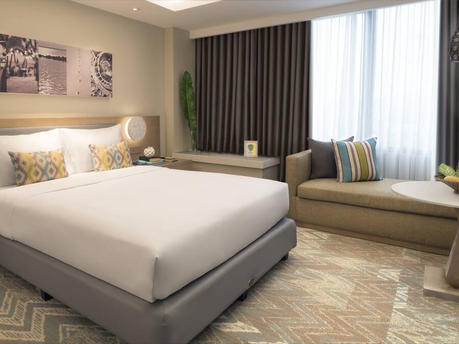 A modern hotel room with a large bed, a sofa, a small table, and a large window with curtains.