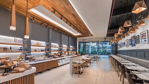 Un café moderno con detalles de madera, una barra larga con exhibición de alimentos y vistas al exterior con vegetación.