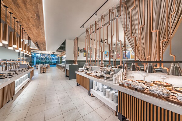 A modern dining area with long communal tables, a buffet setup, and a wooden ceiling.