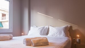 En pænt redt seng med hvide sengetæpper og to sammenrullet håndklæder, et natbord med en lampe, og et vindue med udsigt til en balkon.