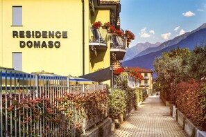 A yellow building with 'RESIDENCE DOMASO' signage, a paved pathway, and a mountainous backdrop. - RESIDENCE DOMASO - ONE BEDROOM APARTMENT (DOMASO)