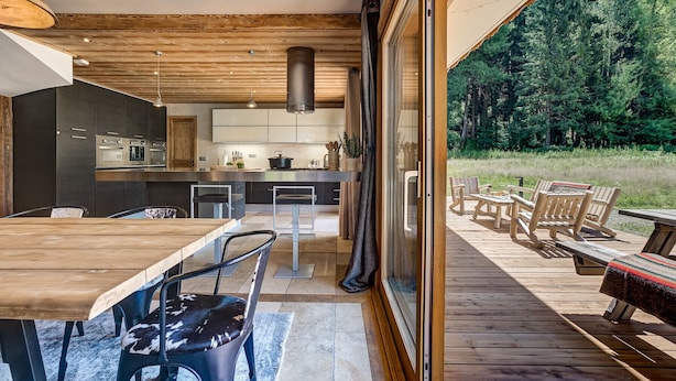 Une cuisine moderne avec des meubles de couleur sombre et des appareils en acier inoxydable, un coin repas avec une table et des chaises en bois, et une terrasse avec des sièges d’extérieur donnant sur une zone boisée.