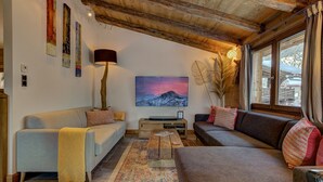 A cozy living room with a wooden ceiling, a large painting of a mountain, and a view of a snowy landscape through the window.