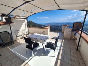 Une terrasse extérieure couverte, dotée d’une table et de chaises, d’un barbecue et offrant une vue sur les montagnes lointaines.