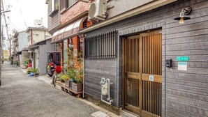 Une rue étroite en milieu urbain, bordée de bâtiments traditionnels japonais, avec une entrée fermée par un portail et un petit espace extérieur agrémenté de plantes.