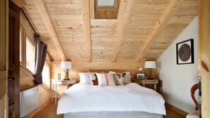 Quarto com teto de madeira, uma cama grande com roupa de cama branca, duas mesas de cabeceira com luminárias e uma pequena mesa com abajur.