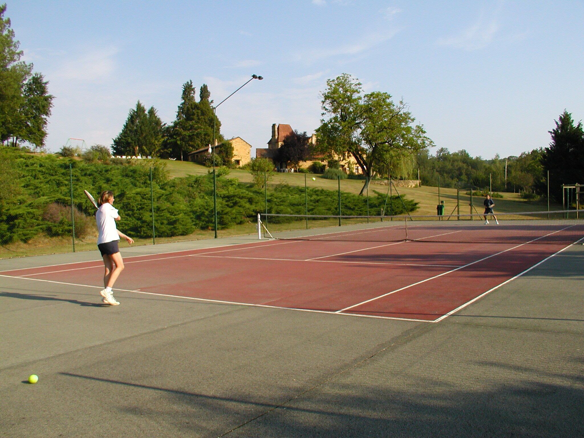 An outdoor tennis court with a net, surrounded by trees and a building in the background.