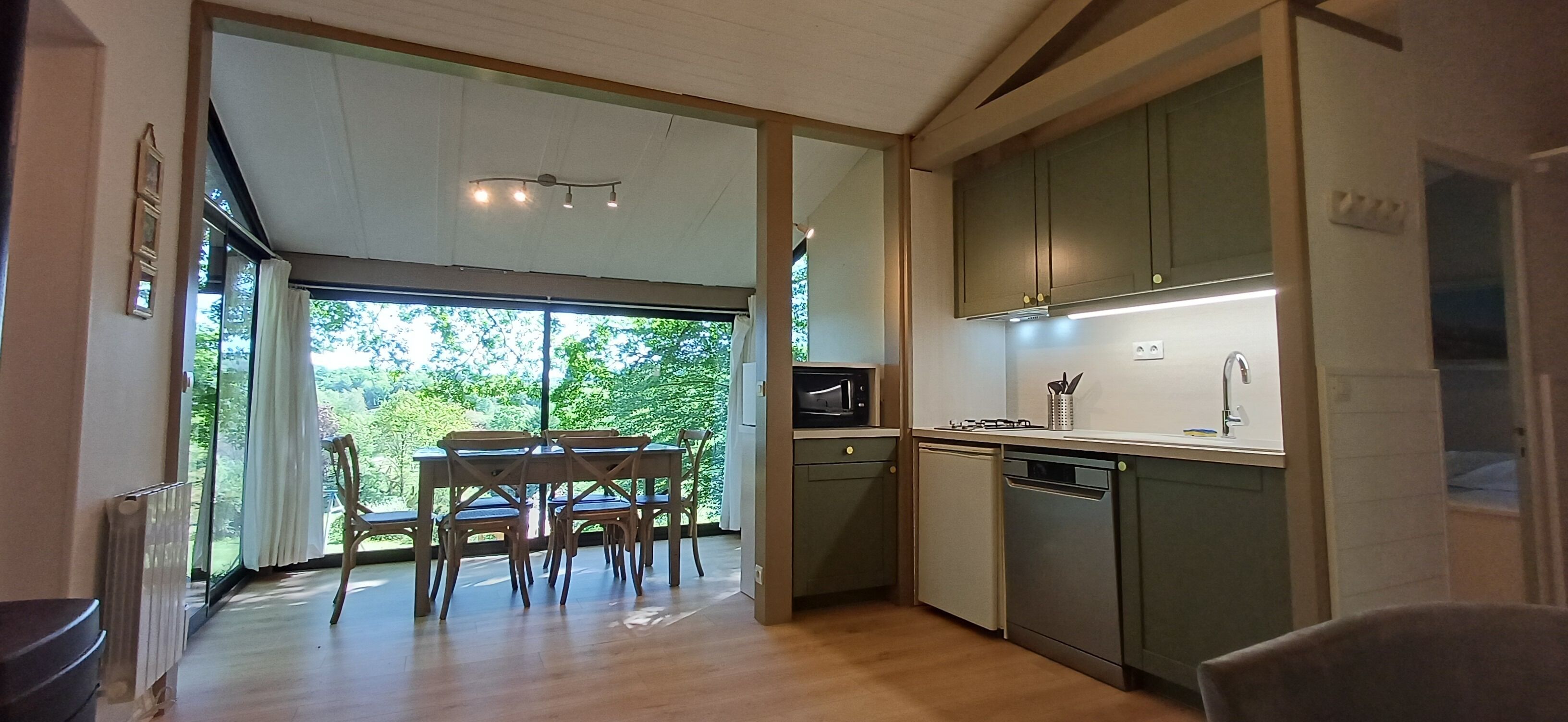 A modern kitchen with wooden cabinets, a dining area with a table and chairs, and a view of greenery outside.
