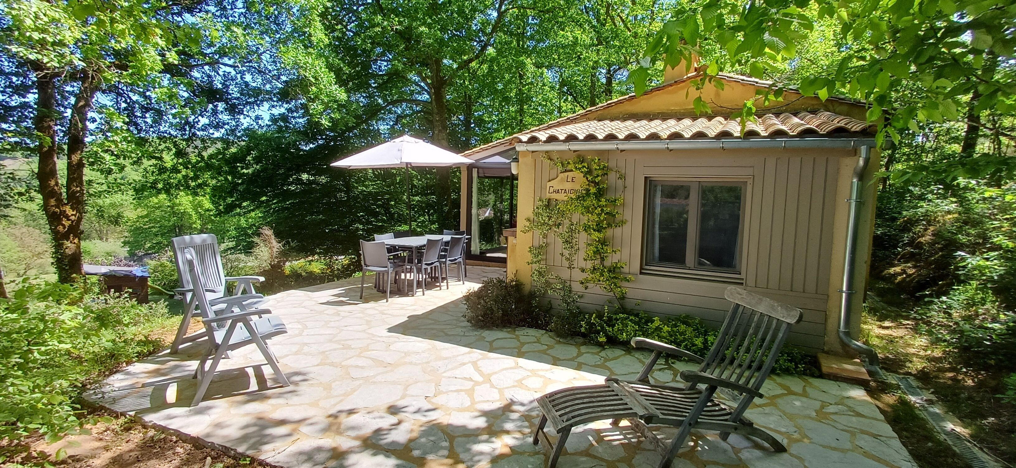 Un petit bâtiment d’un seul étage, avec un toit en tuiles et une enseigne indiquant « LE CHATEAUMONT ». Il comprend une terrasse avec un sol en pierre, du mobilier d’extérieur et une végétation luxuriante qui entoure les lieux.