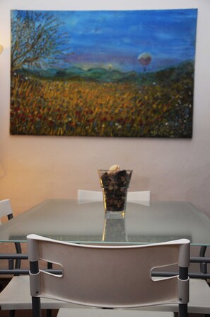 Una mesa de comedor con superficie de vidrio, rodeada de sillas, y un cuadro de un paisaje con un globo aerostático en la pared de fondo.