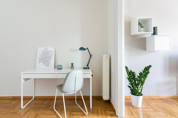 Un bureau minimaliste composé d’un bureau blanc, d’une chaise, d’une carte encadrée, d’une lampe de bureau, d’une petite plante et d’un radiateur.
