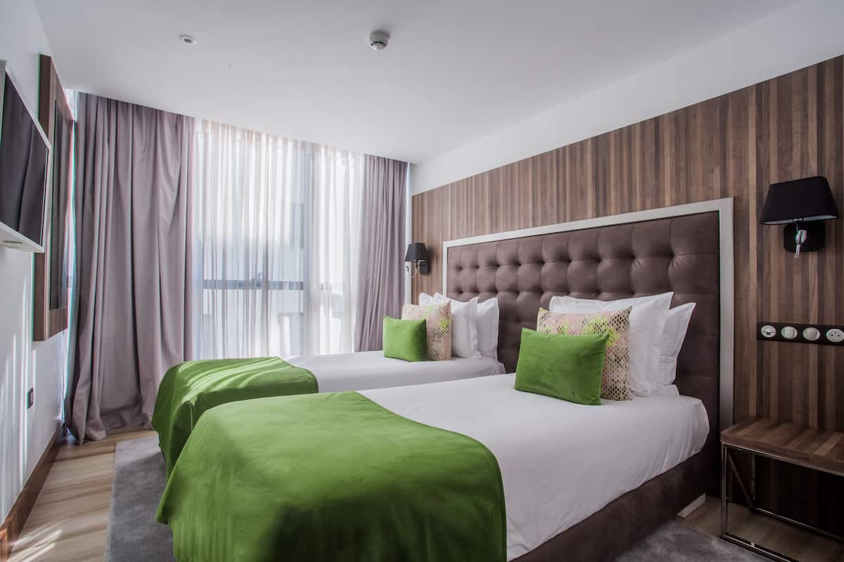 A hotel room with two beds, a large window with curtains, a flat-screen TV, and a wall-mounted lamp.