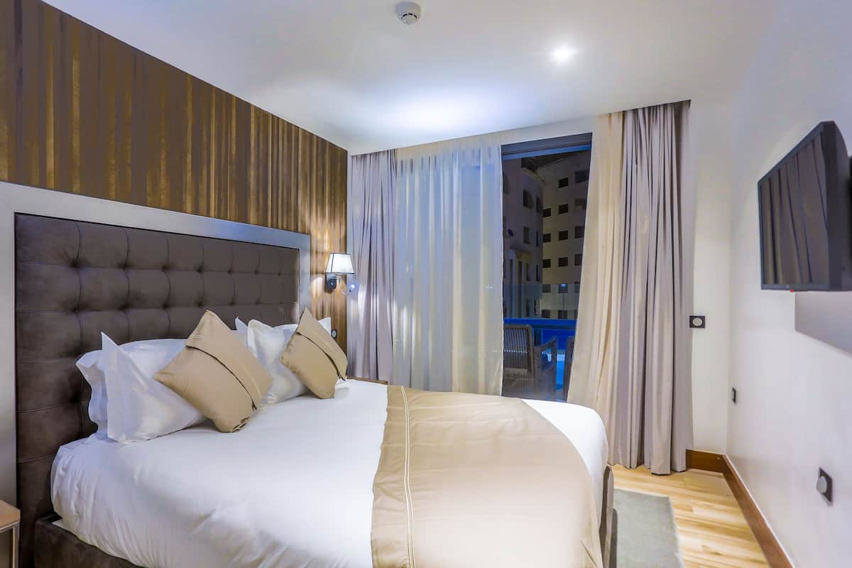 A hotel room with a large bed, a headboard, a bedside table, a lamp, and a view of a building through the window.