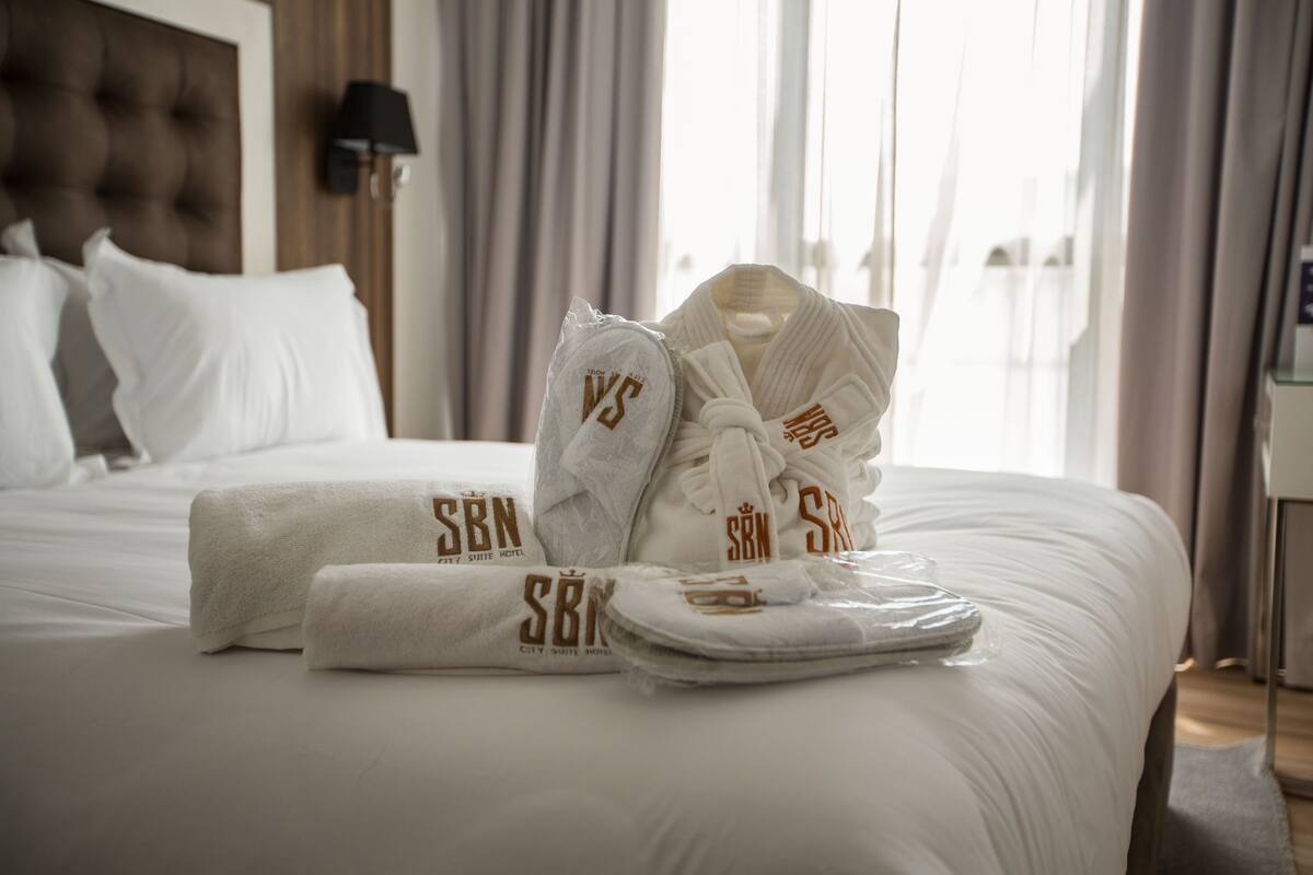 A neatly made bed with white linens and a set of towels and robes with 'SBN' branding.