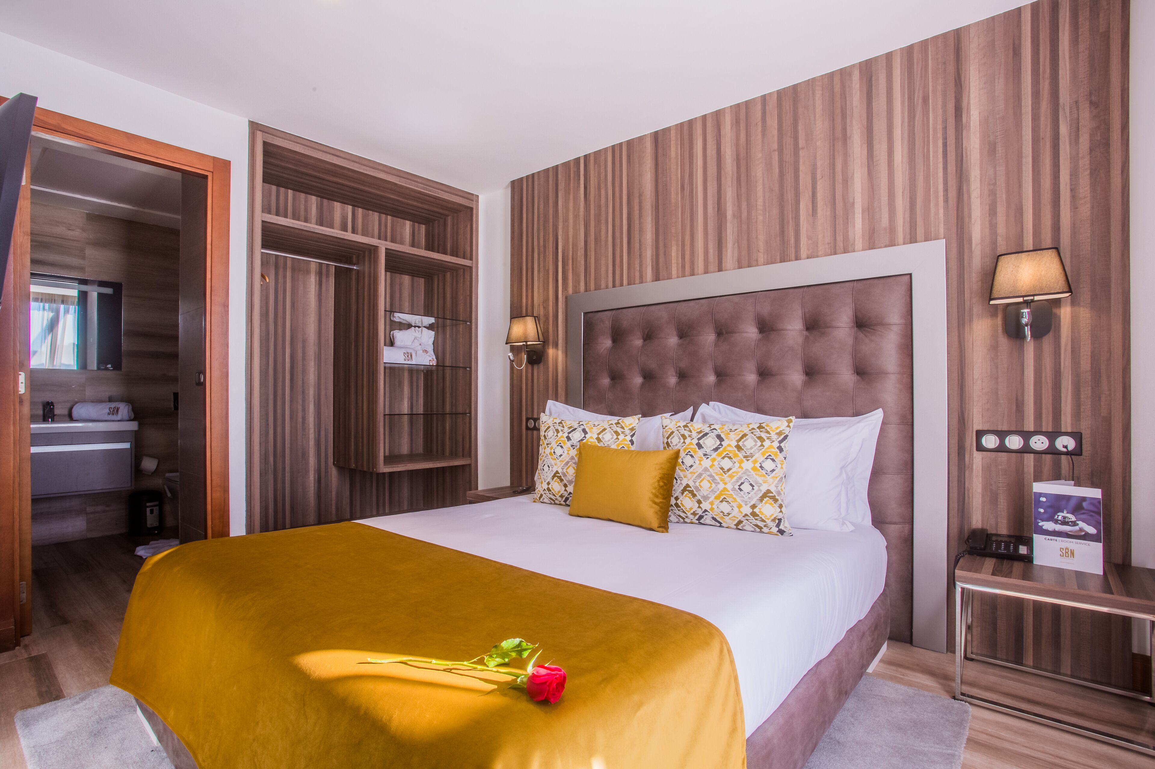 Habitación de hotel con una cama grande, paneles de madera, una mesita de noche con lámpara y un baño visible a través de una puerta abierta.