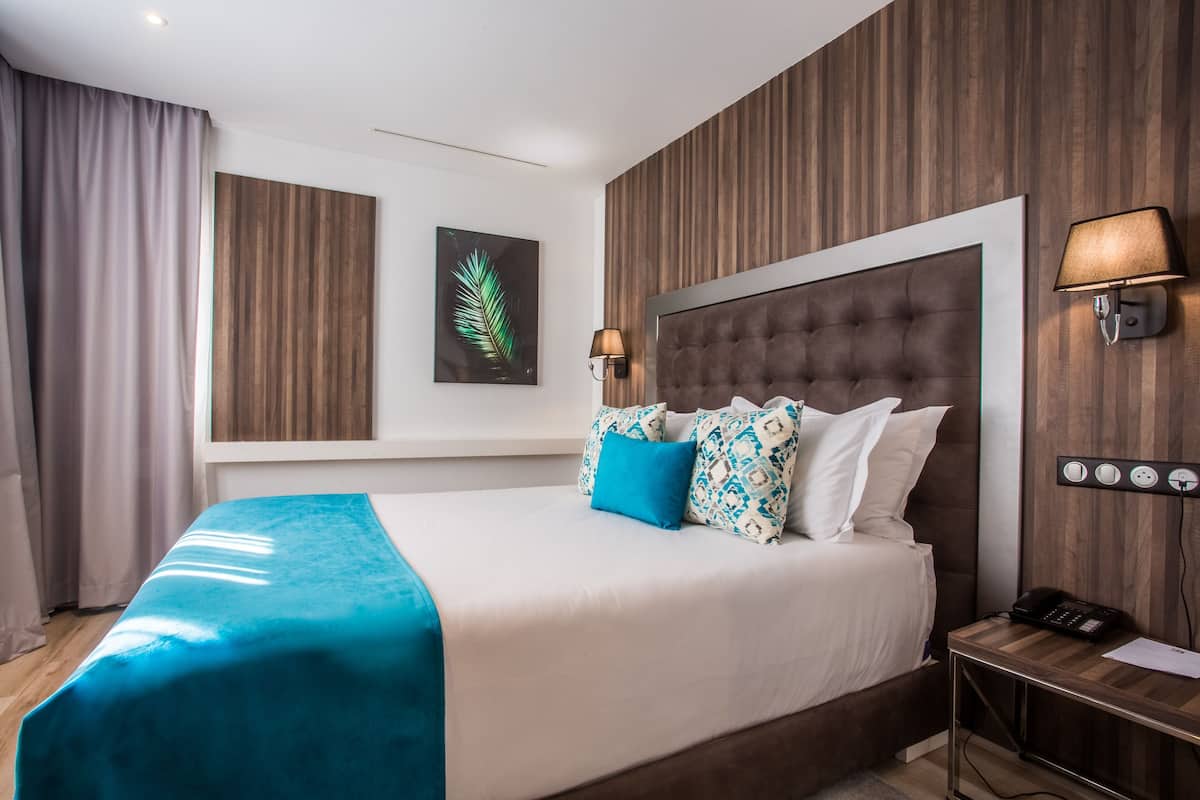 A hotel room with a large bed, a nightstand, a lamp, a phone, and a wall-mounted light switch.