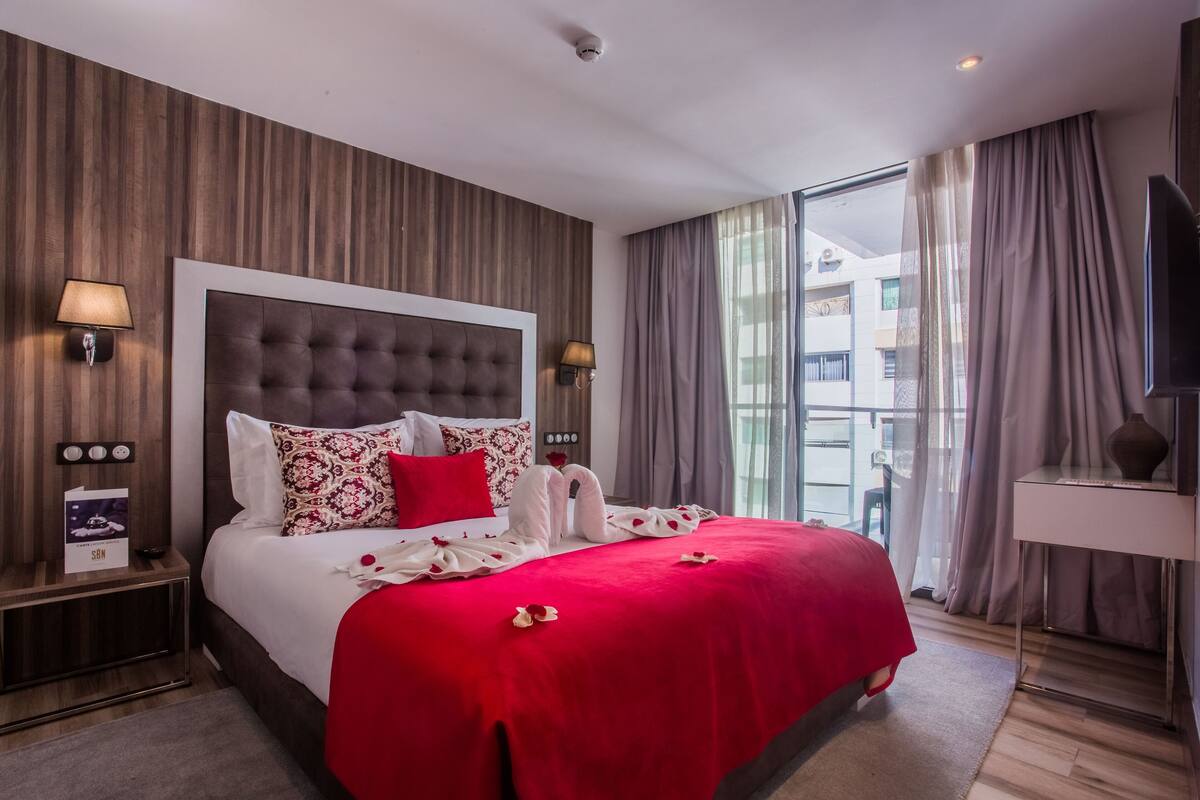 A hotel room with a large bed, a headboard, two wall-mounted lamps, a nightstand with a lamp, a TV, and a view of the outside through large windows.