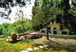Un bâtiment en pierre doté d’une cheminée en pierre et d’une table de pique-nique en bois, le tout entouré d’arbres.