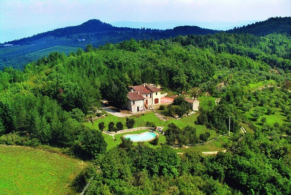 Une maison en pierre, surmontée d’un toit de tuiles rouges, est entourée d’une végétation luxuriante et se dresse sur un arrière-plan de collines.