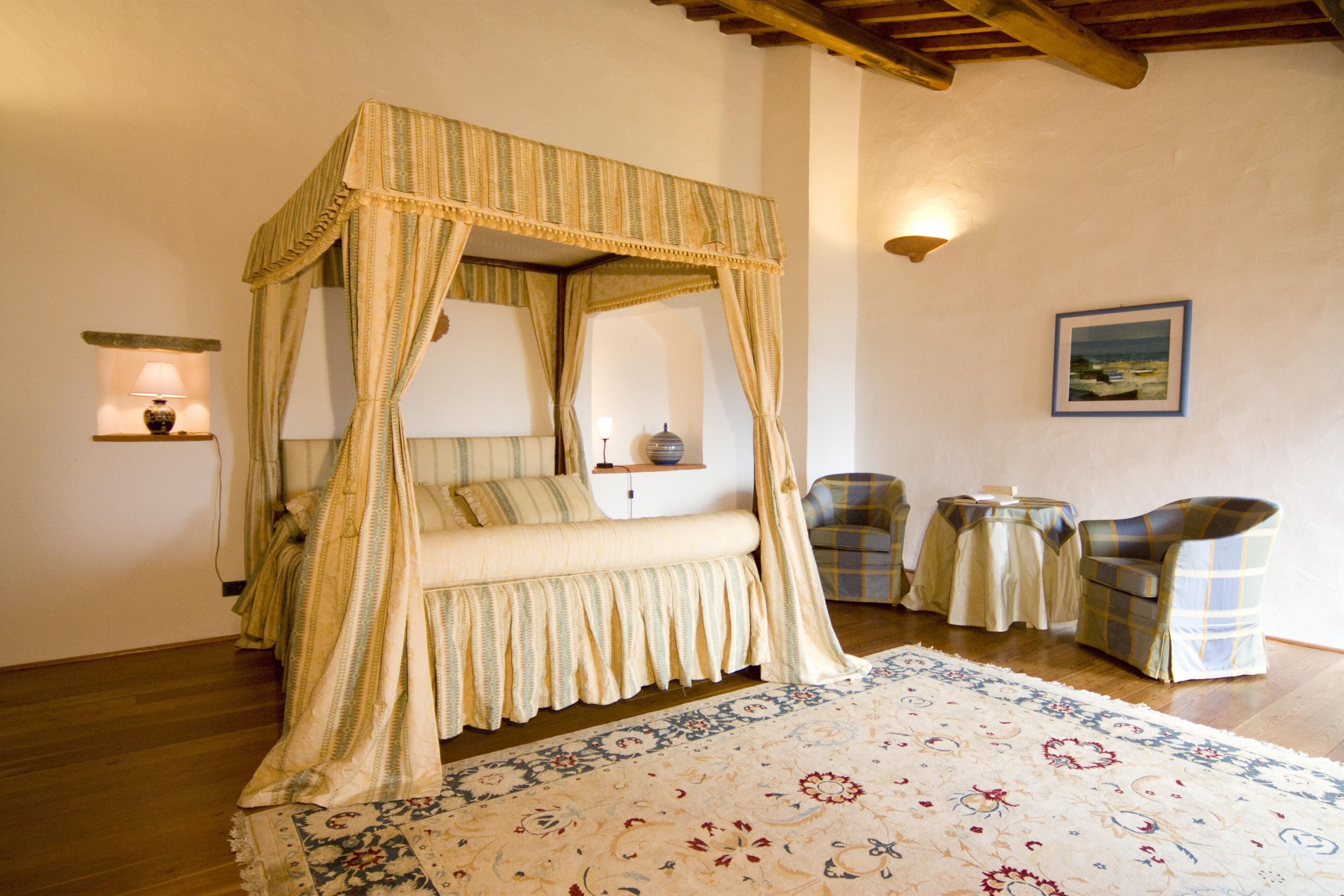 A four-poster canopy bed with a floral-patterned rug, two armchairs, and a small table with a lamp.