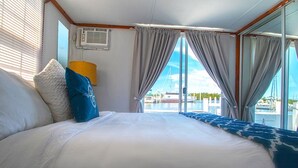 Uma cama grande com colcha estampada em branco e azul, um travesseiro azul e um travesseiro branco. Há uma janela com cortinas cinzas, um abajur amarelo e vista para um porto com barcos e edifícios.