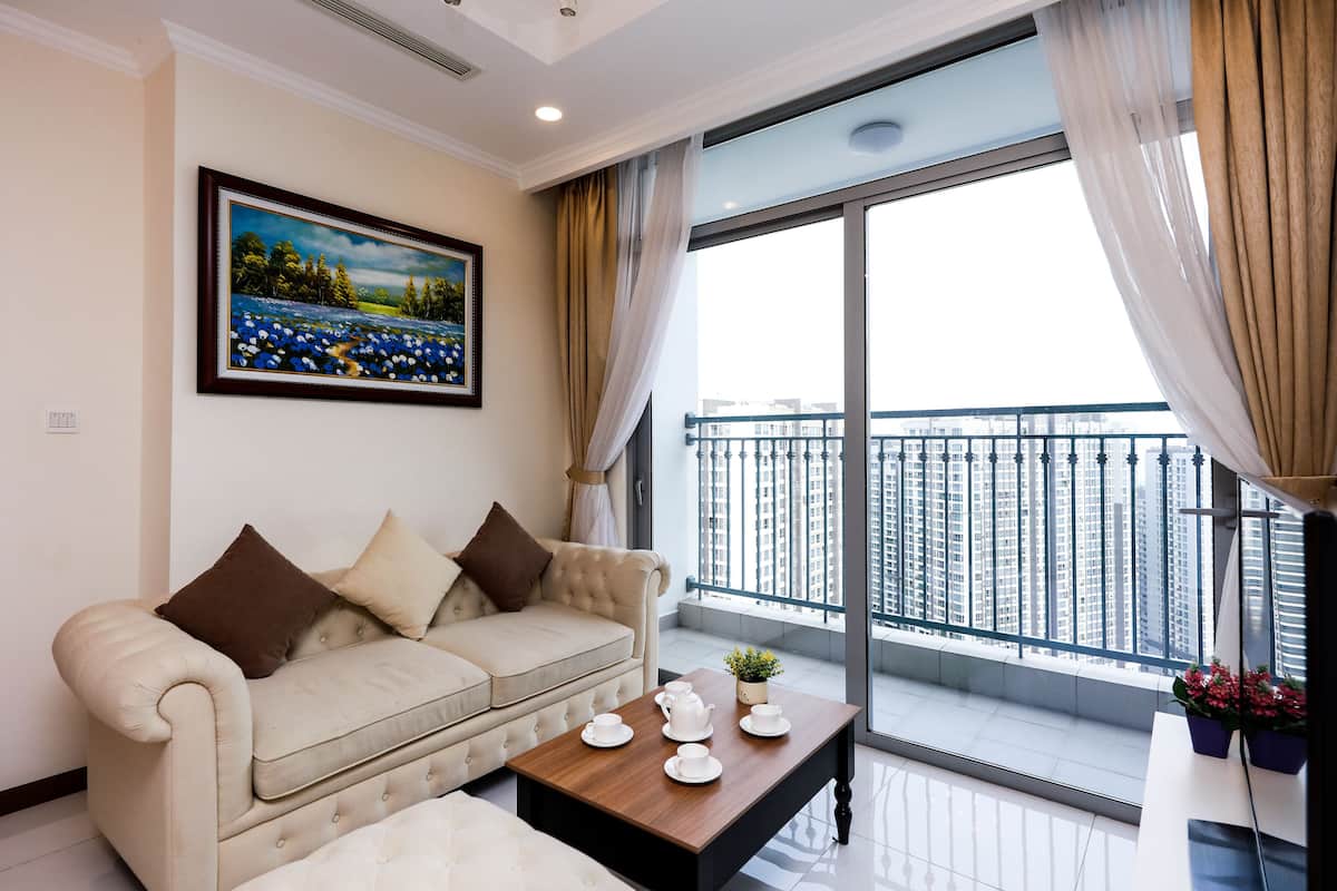 Een moderne woonkamer met een bank, een salontafel met theeservies, een schilderij aan de muur, en een balkon met uitzicht op hoge gebouwen.
