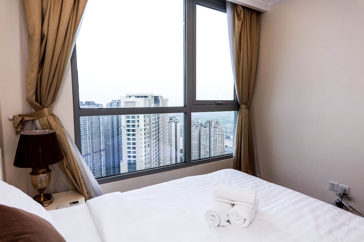 Een hotelkamer met een groot raam en uitzicht over de stad, een net opgemaakt bed en een nachtkastje met een lamp.