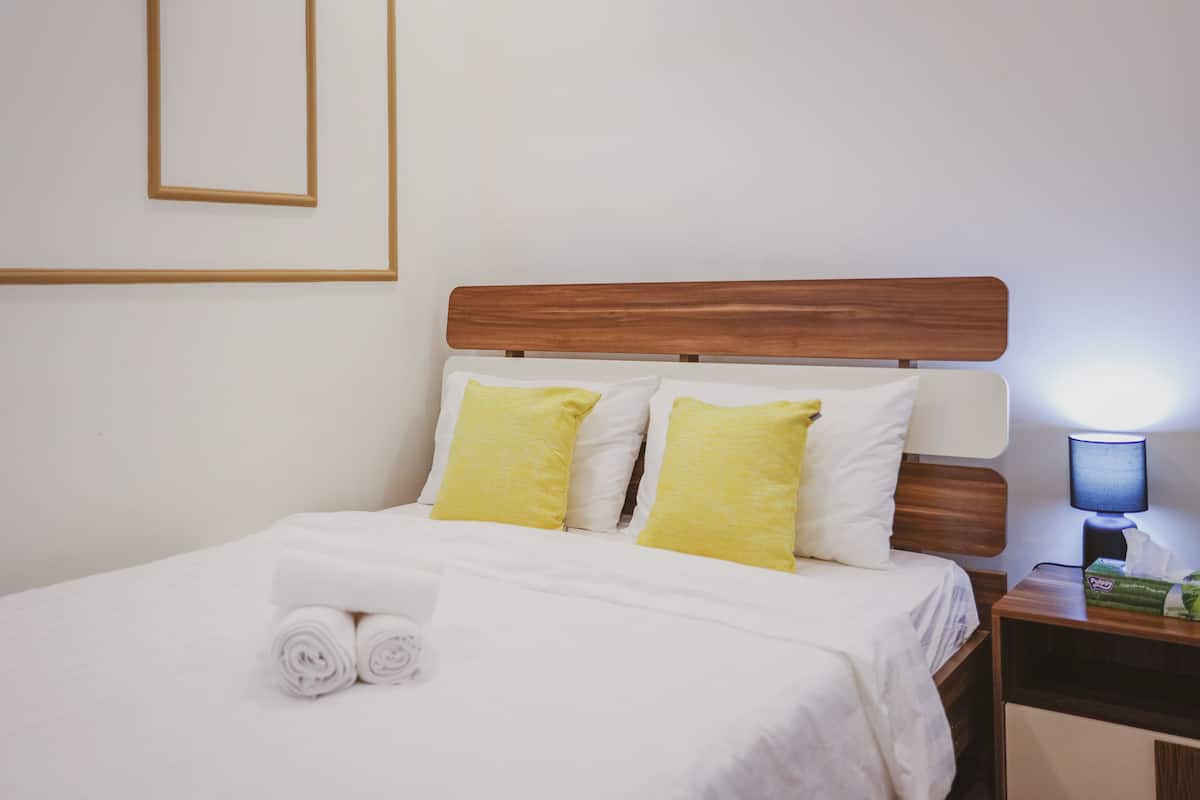 Een netjes opgemaakt bed met witte lakens en twee gele kussens, een houten hoofdbord en een nachtkastje met een lamp en zakdoekjes.